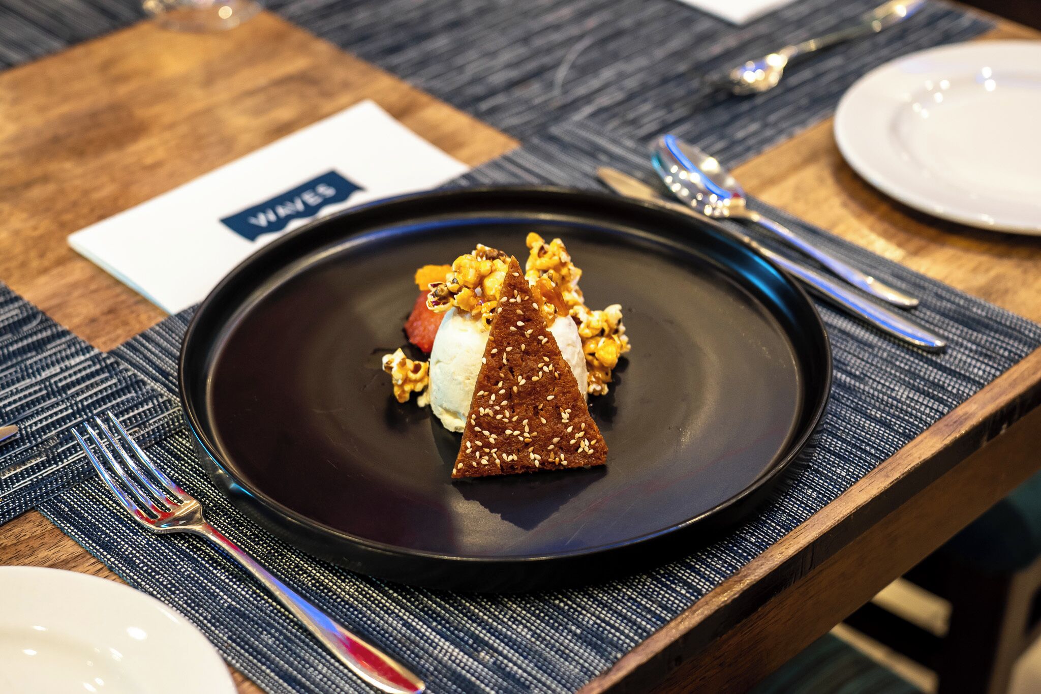 Ein Desserteller mit einem dreieckigen Kuchenstück, Eiscreme und karamellisierten Nüssen auf einer schwarzen Tellermitte.