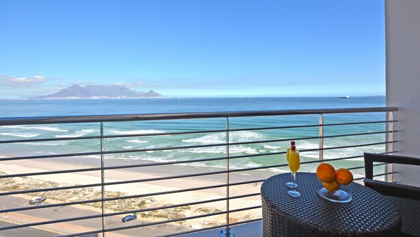 Een balkon met uitzicht op de oceaan, een tafel met drinken en fruit, en een stoel.