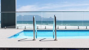 Een zwembad met helderblauw water, een glazen balustrade en uitzicht op de oceaan en verre bergen.