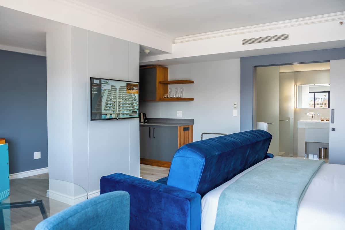 Een moderne hotelkamer met een groot bed, een blauwe bank, een kitchenette en een zichtbare badkamer door een open deur.
