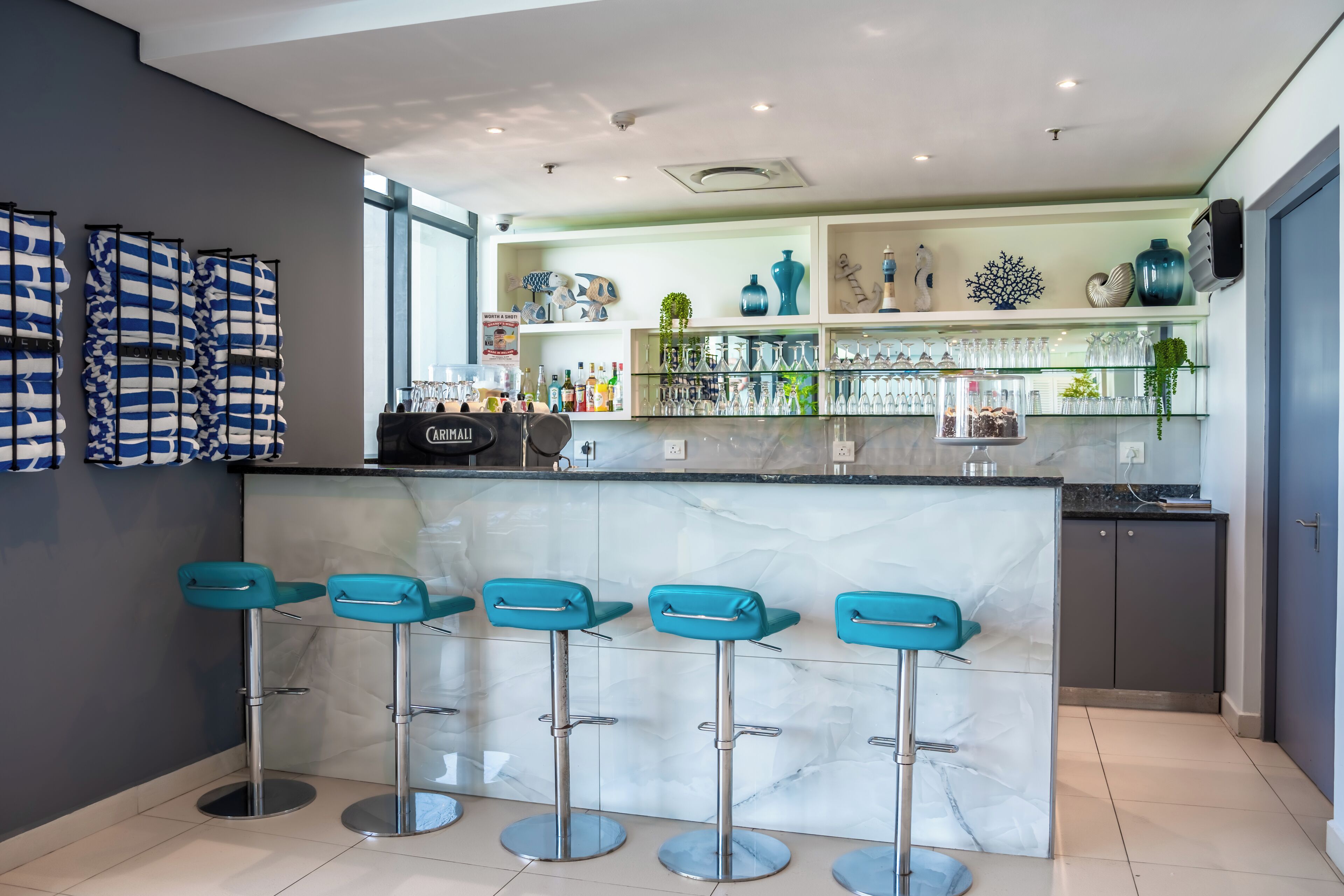 A modern bar area with a marble countertop, teal bar stools, and glass shelves displaying bottles and decorative items.