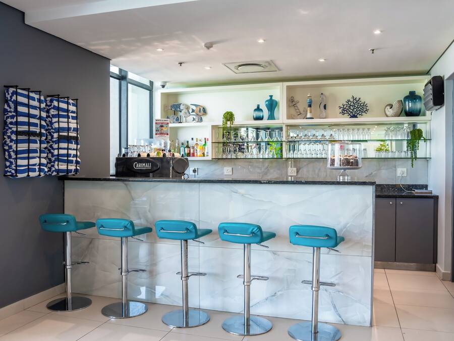 A modern bar area with a marble countertop, teal bar stools, and glass shelves displaying bottles and decorative items.