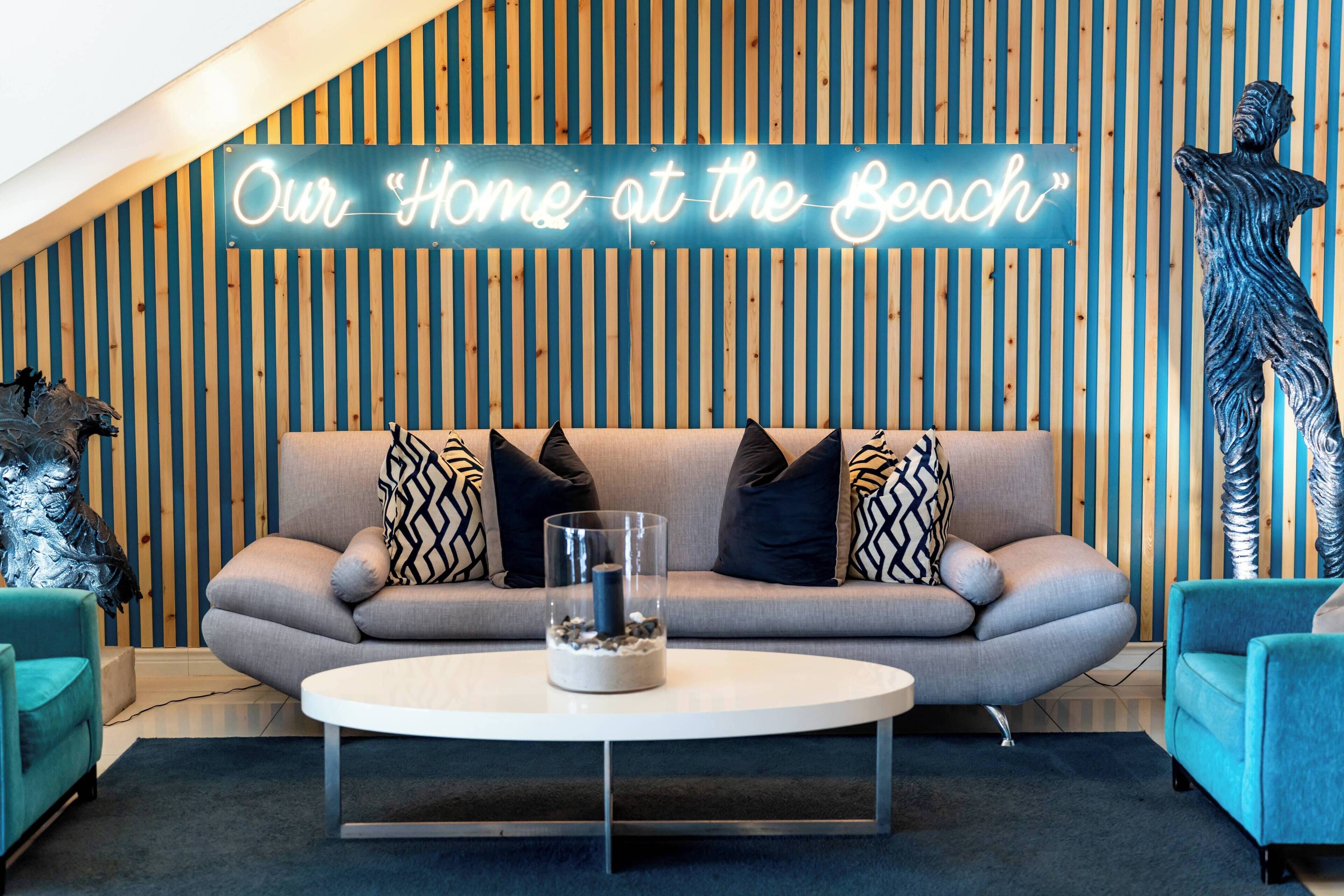 Un salon moderne avec un canapé, une table basse en verre et un panneau lumineux néon sur lequel on peut lire : « Notre maison au bord de la mer ».