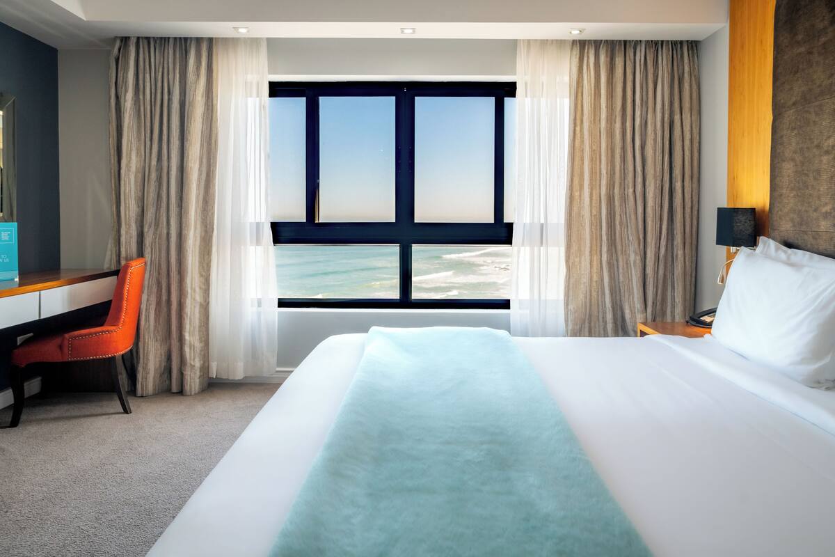 Een hotelkamer met een groot bed, een bureau en een stoel. Er is uitzicht op de oceaan door een groot raam.