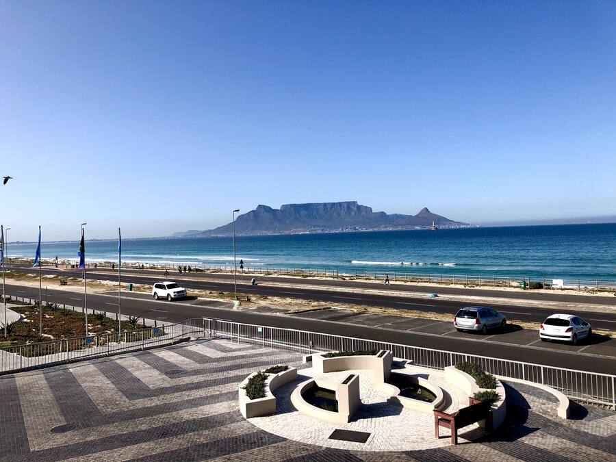 A coastal road with a pedestrian walkway, a parking area, and a view of the ocean and a mountain in the distance.