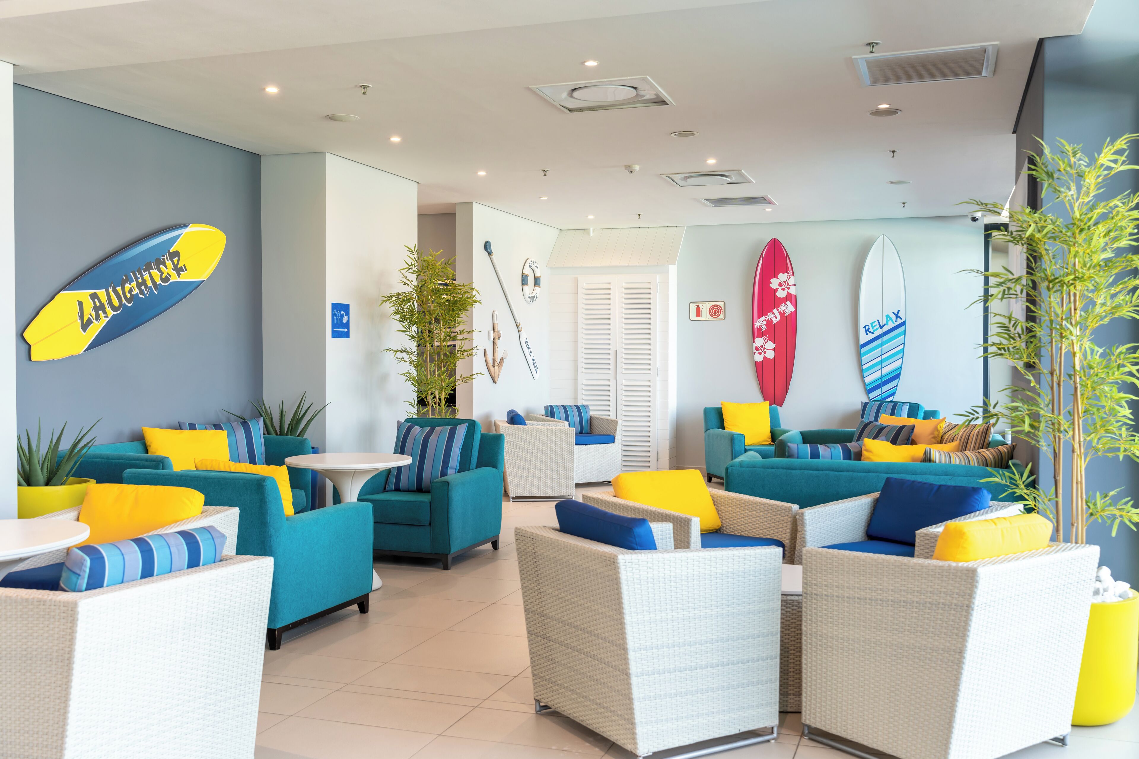 A modern lounge area with wicker furniture, yellow and blue cushions, and surfboards on the wall.