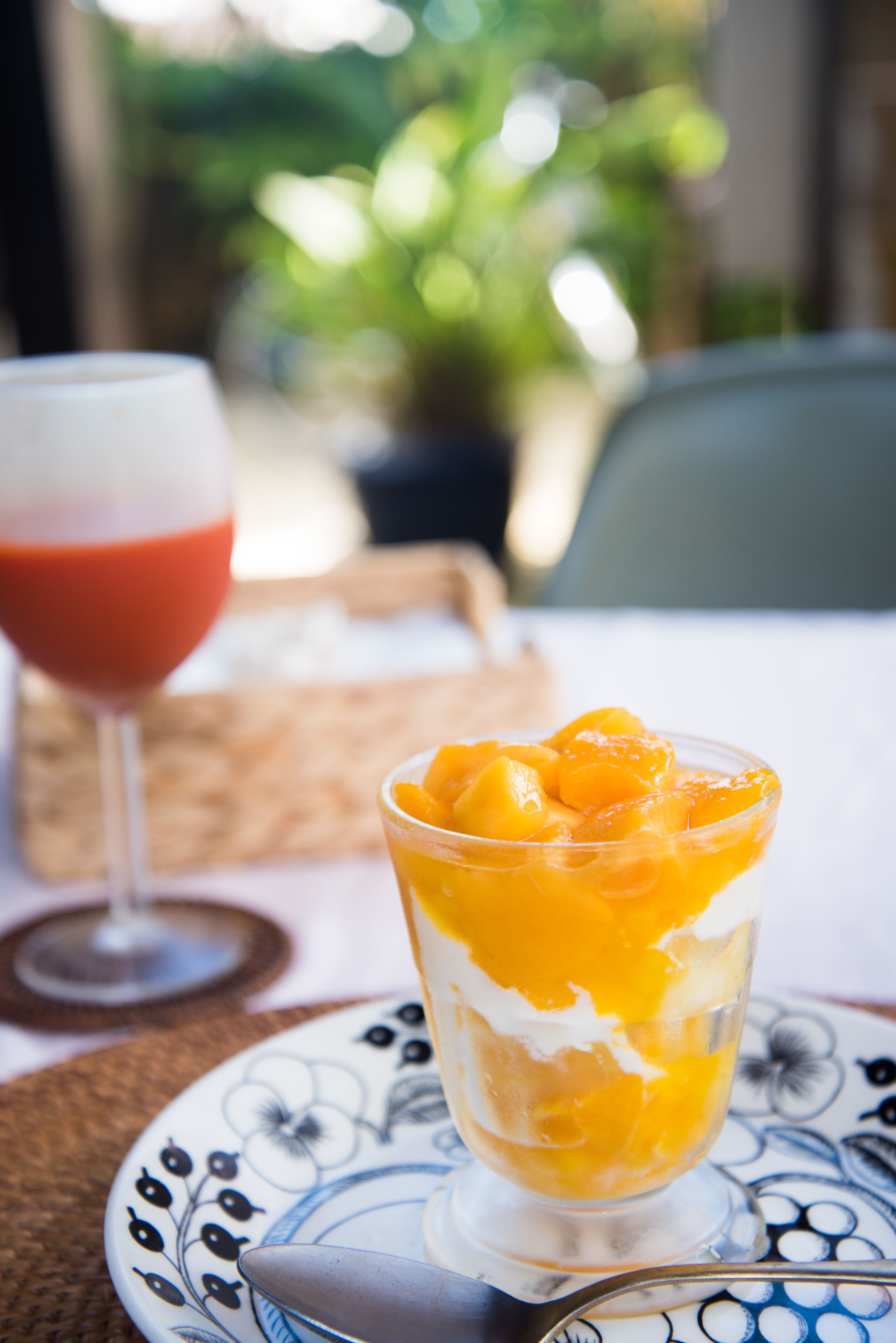 A glass of orange juice with peach chunks on a patterned plate, a glass of red drink, and a wicker placemat.