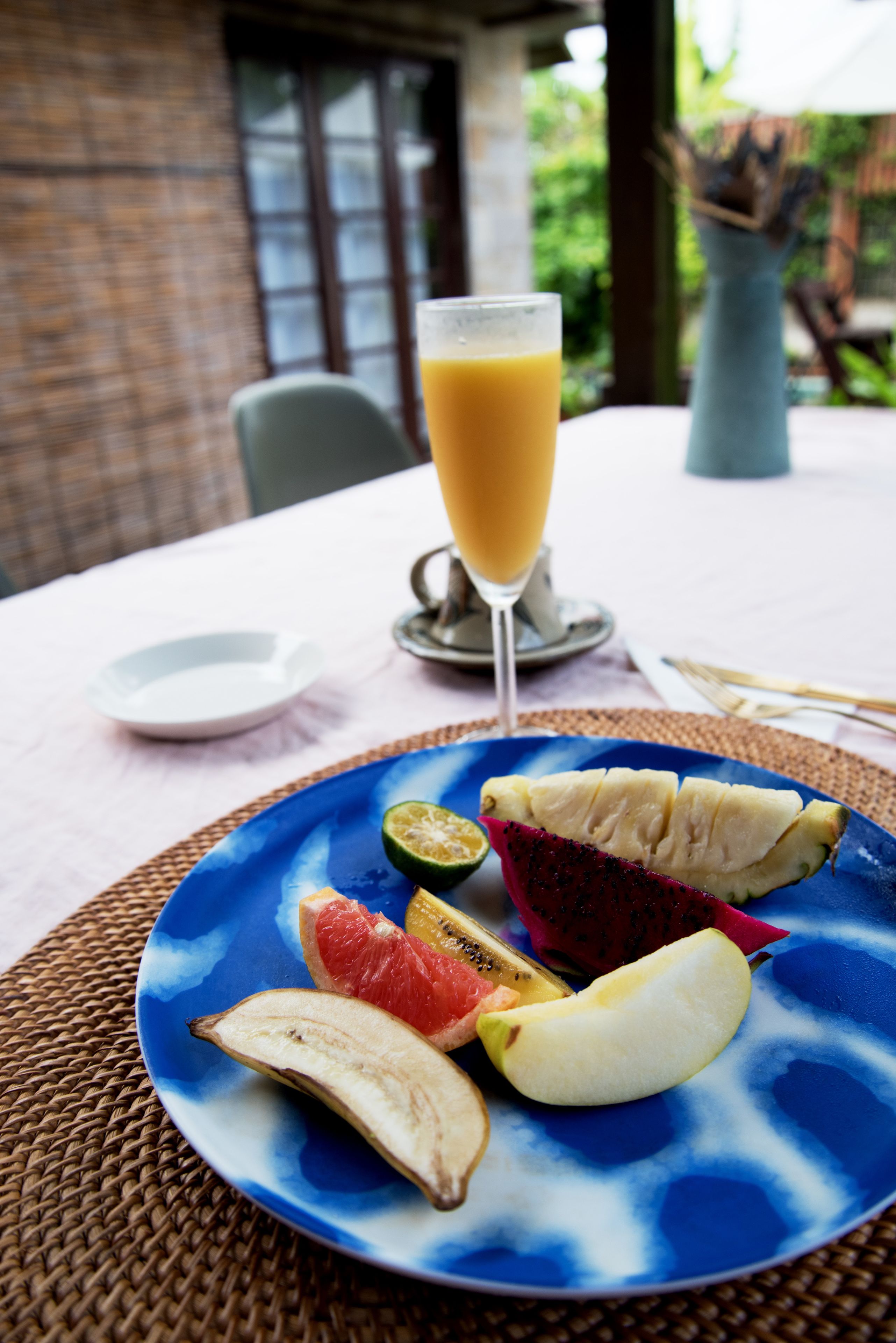 Une table avec une assiette bleue remplie de fruits, un verre de jus d’orange et une assiette blanche.