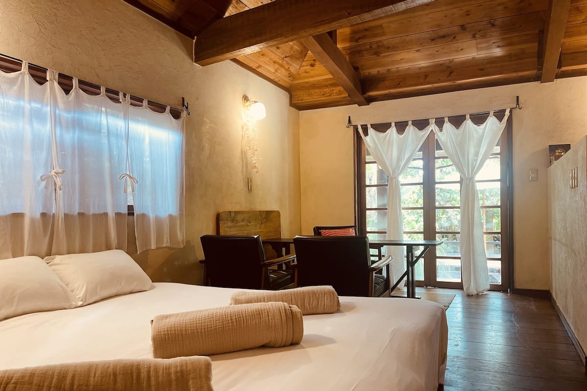 A bedroom with a large bed, a wooden ceiling, a desk with a chair, and a window with curtains.