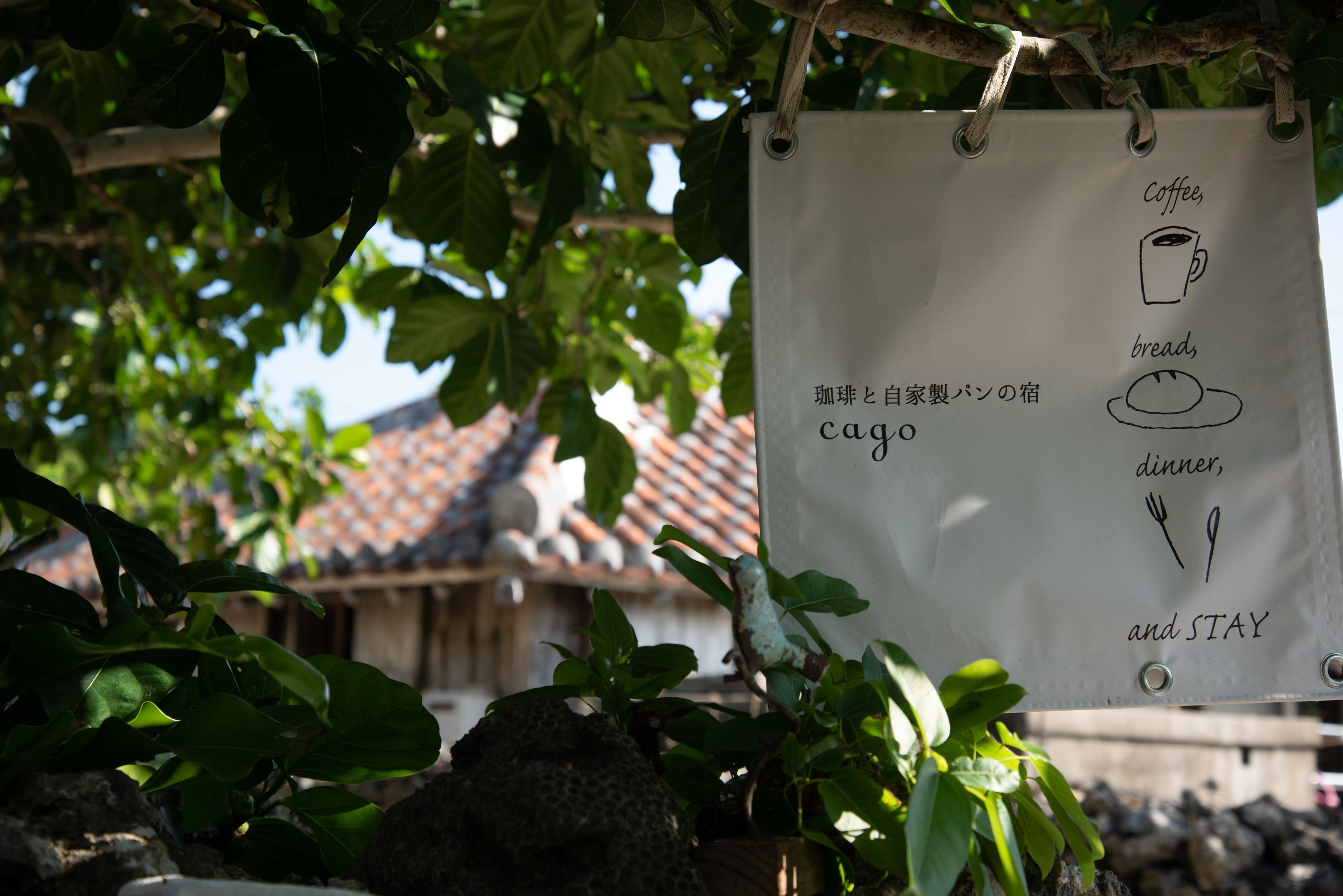 Un letrero colgando de un árbol con texto e ilustraciones de una taza de café, pan, cubiertos y las palabras 'cago' y 'STAY'.