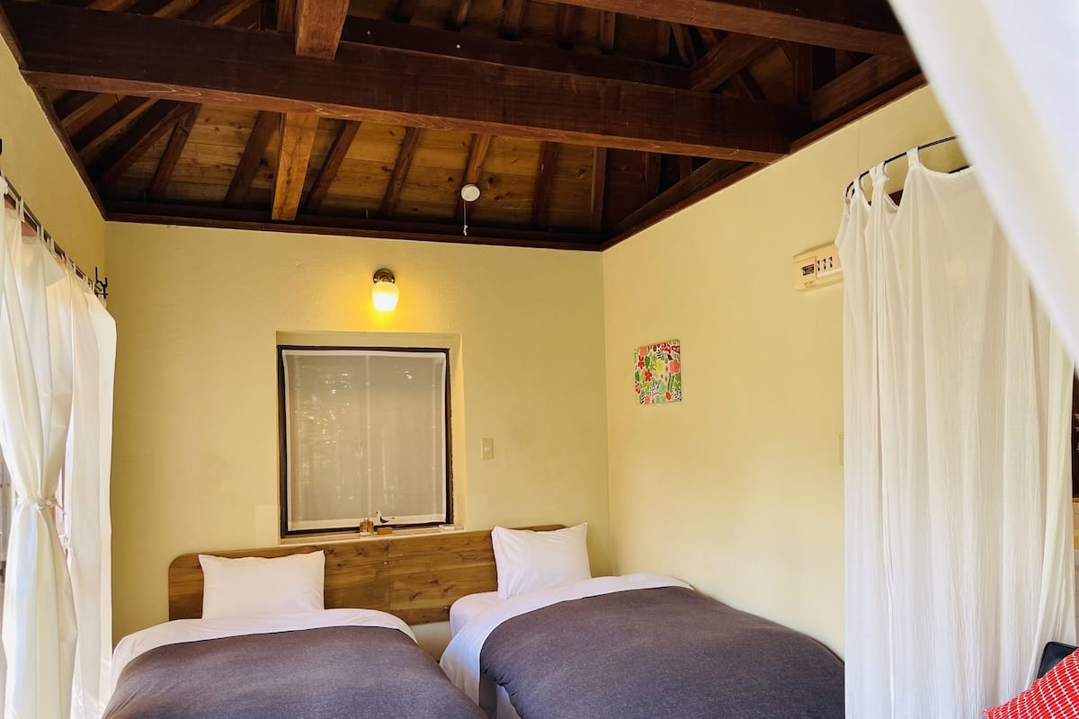 A room with two beds, wooden headboard, a window, a wall clock, and a bed with a red and white pillow.