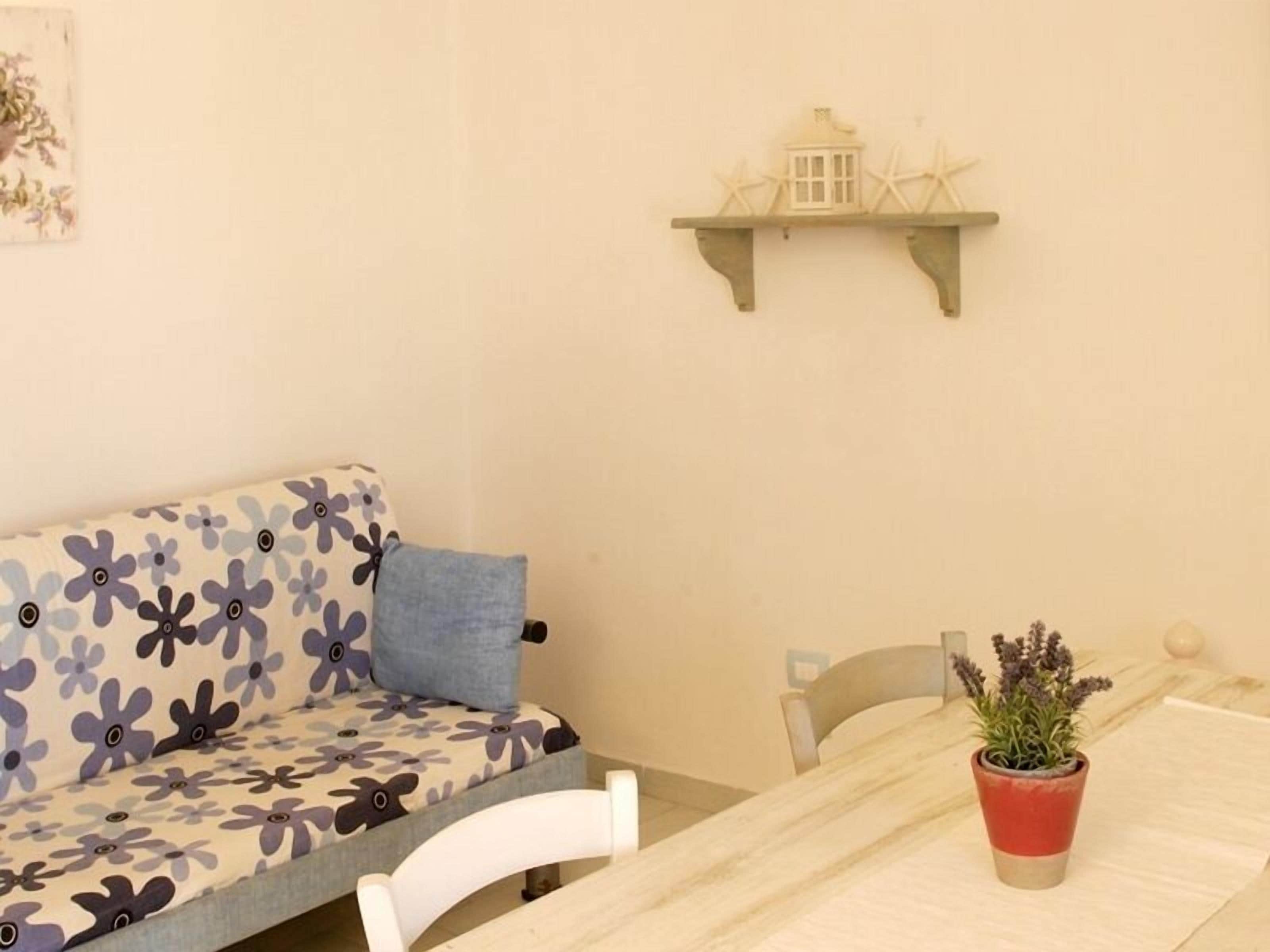 Una habitación acogedora con un sofá floral, una mesita con una planta en maceta y un estante de pared con un modelo de faro.