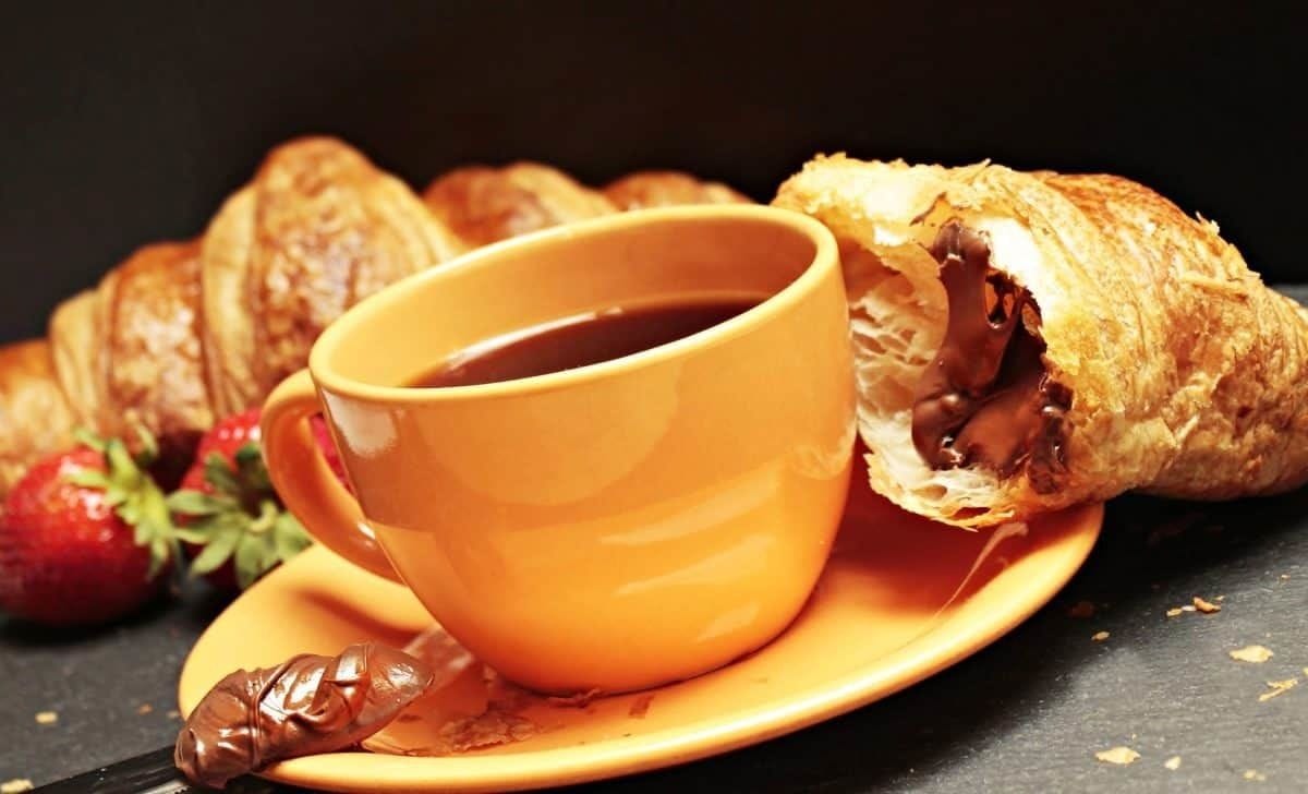 A cup of coffee on a saucer with a croissant and a chocolate-covered item on a plate.