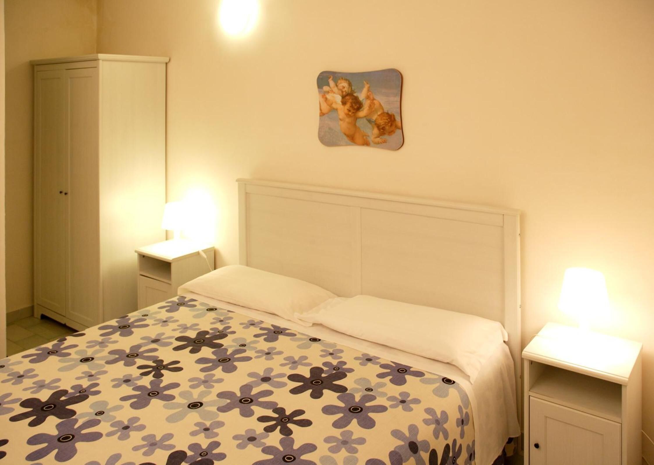 Um quarto com uma cama, duas mesas de cabeceira, um criado-mudo e um guarda-roupa, além de um quadro na parede.