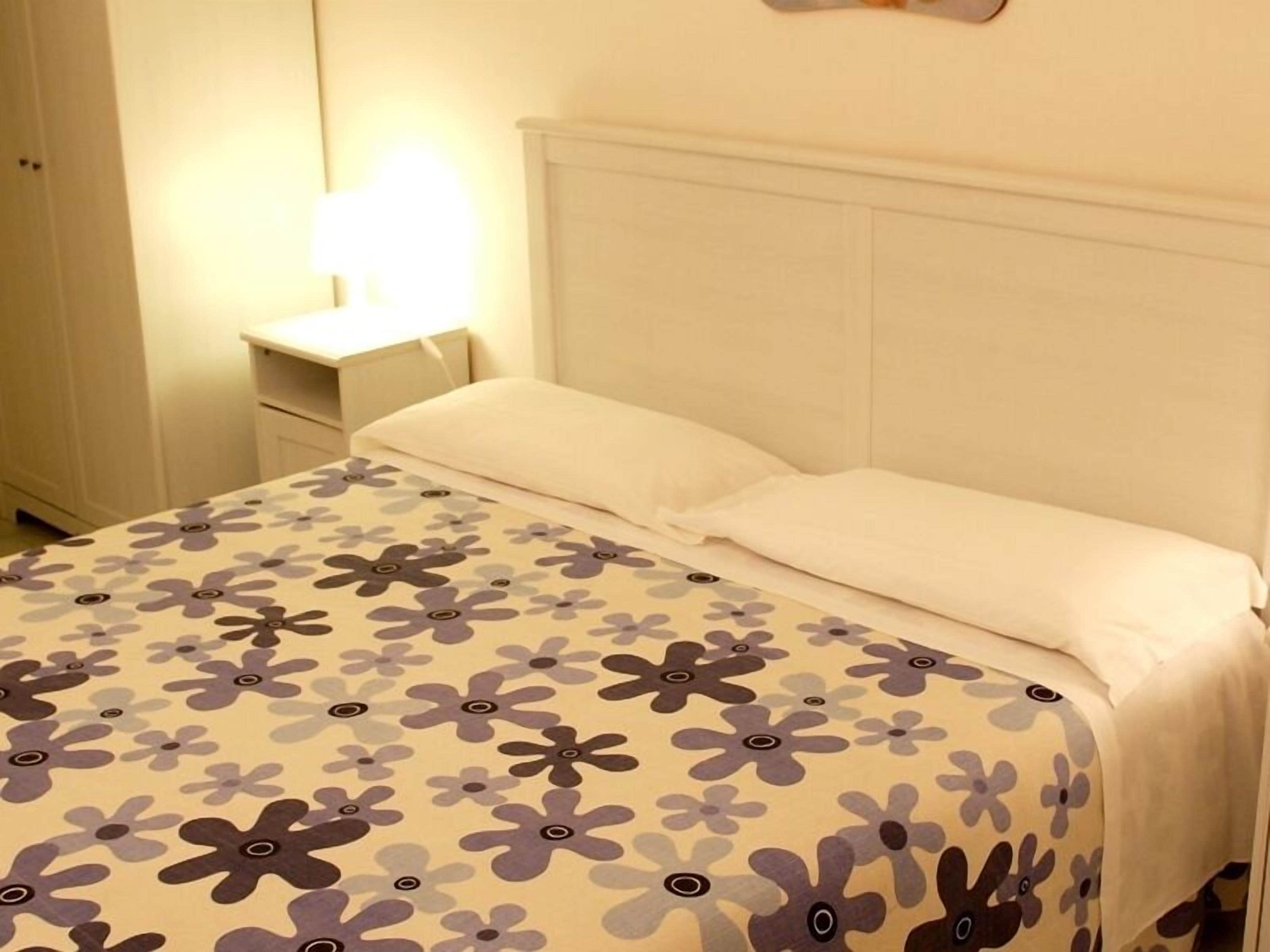 Un lit bien fait, recouvert d’une couvre-lit à motifs floraux et agrémenté d’oreillers blancs, dans une chambre dont la tête de lit est blanche et qui comprend une table de chevet avec une lampe.