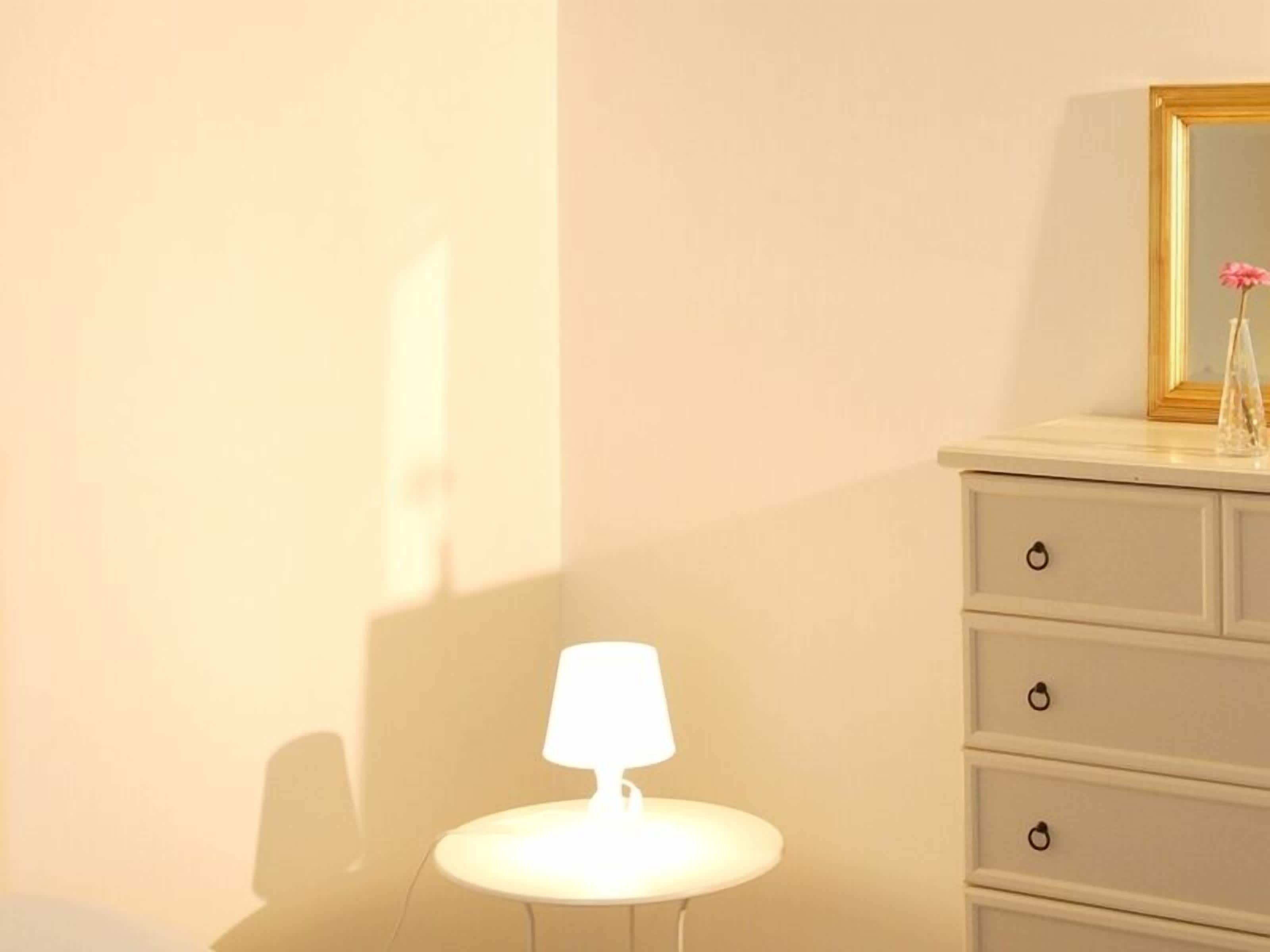 A room with a bedside lamp, a dresser with drawers, and a mirror with a gold frame.