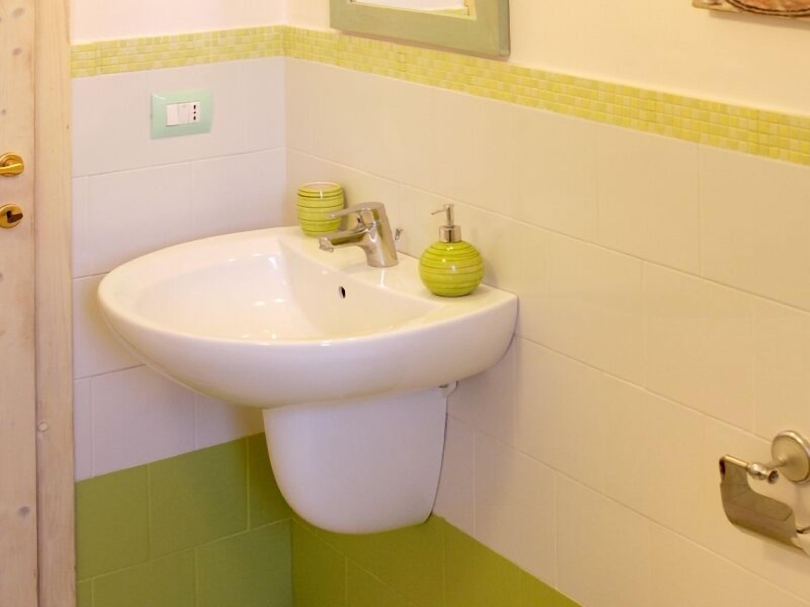 A bathroom with a white sink, green and yellow tiled walls, a green soap dispenser, and a light switch on the wall.