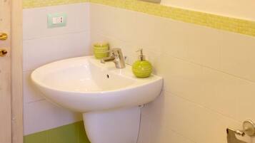 Une salle de bain avec un lavabo blanc, des murs recouverts de carreaux verts et jaunes, un distributeur de savon vert et un interrupteur sur le mur.