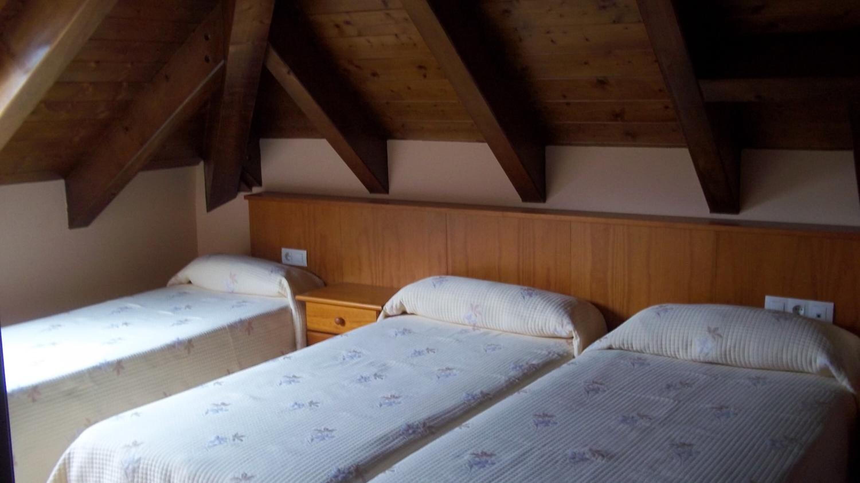 A room with three single beds aligned against a wooden wall, each with a wooden headboard and a small bedside table.