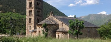 Una iglesia de piedra con un campanario y una aguja, rodeada de abundante vegetación y montañas.
