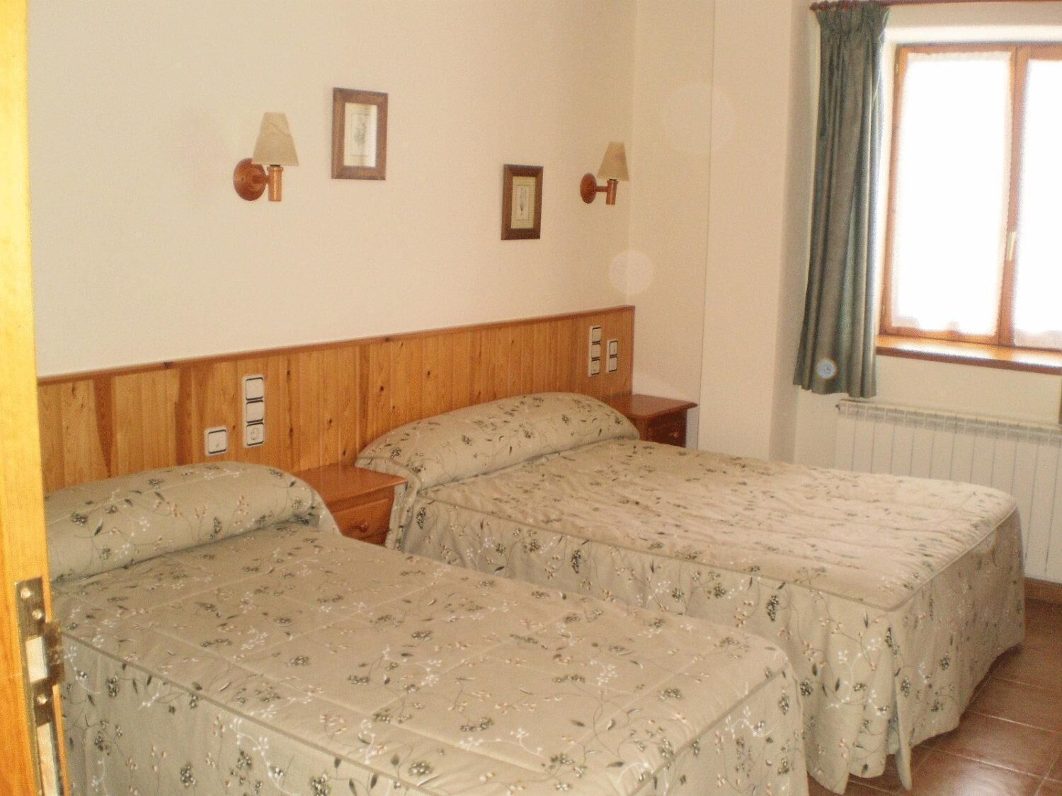 A room with two beds, wooden headboards, wall-mounted lamps, and a window with curtains.