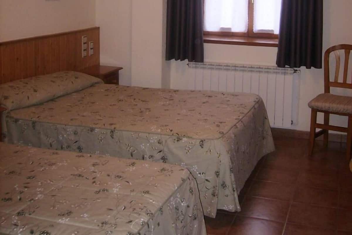 Una habitación con dos camas, una silla, una ventana con cortinas, una lámpara de pared y un radiador.