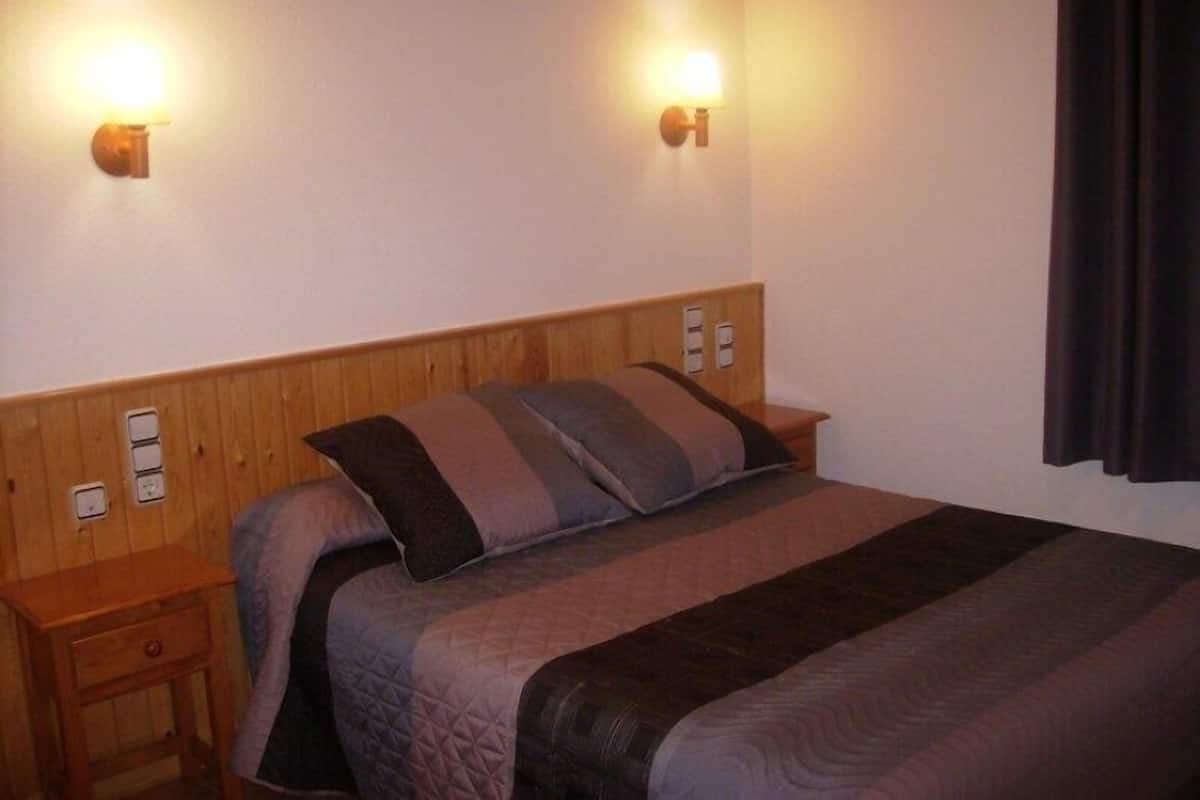 Habitación de hotel con cabecera de madera, cama con cubrecama acolchado, dos mesitas de noche y lámparas fijadas a la pared.