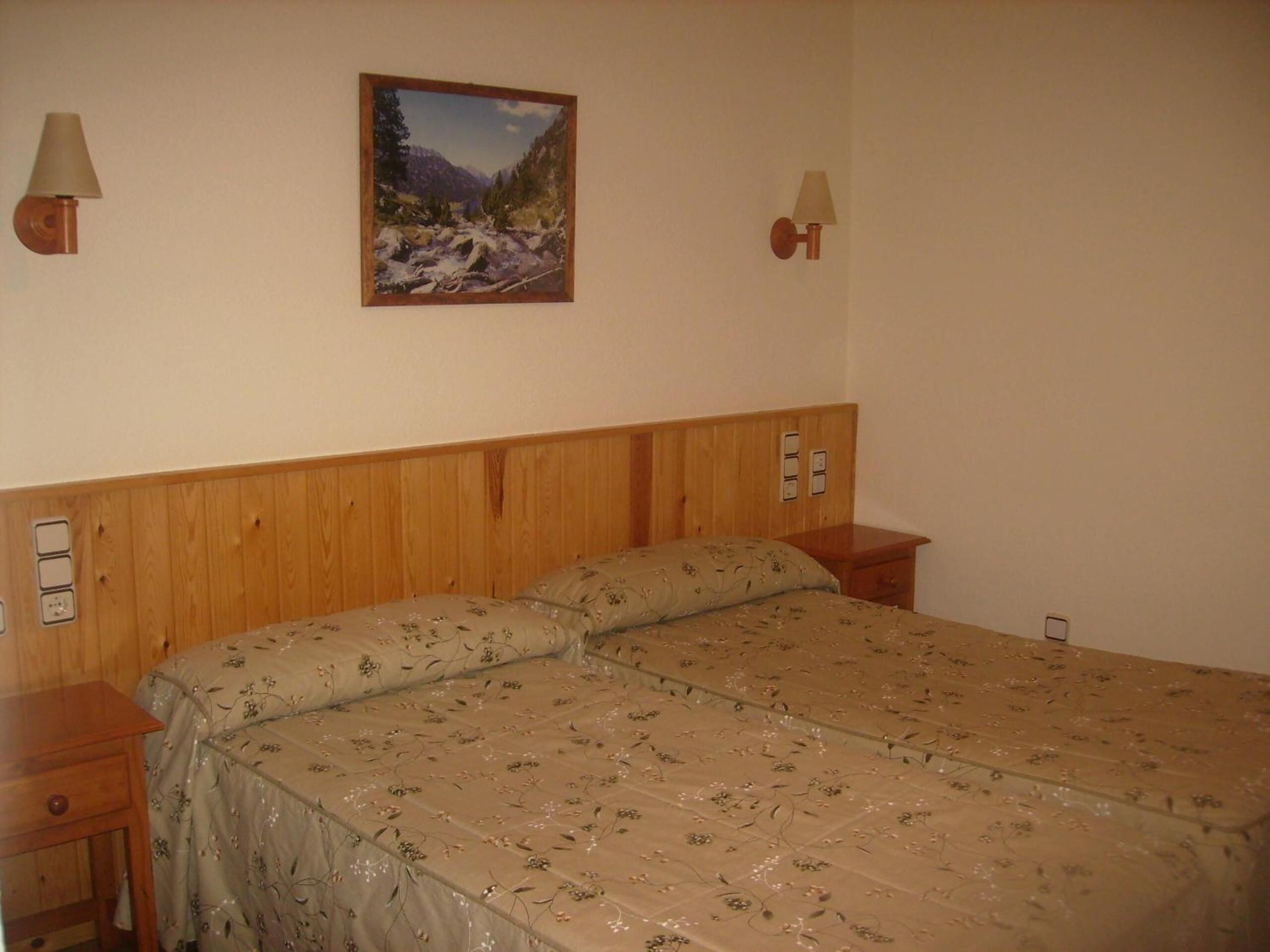 Un dormitorio con cabecera de madera, dos mesitas de noche, una cama con ropa de cama floral, un cuadro de un paisaje montañoso y dos lámparas de pared.