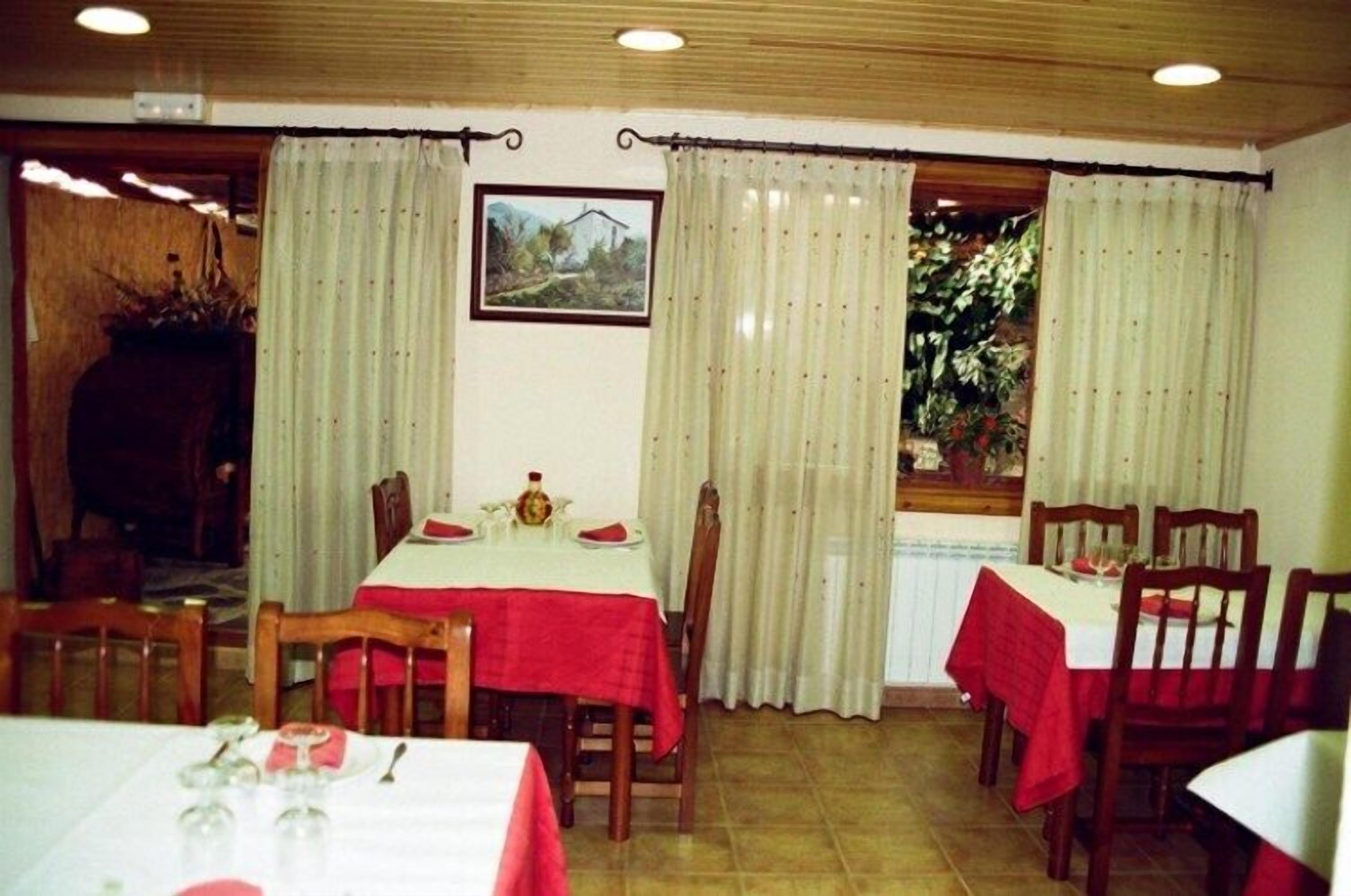 A dining area with tables set for four, each with a red tablecloth and white tablecloths. There are curtains on the windows, and a framed picture on the wall.