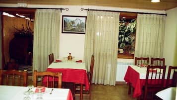 A dining area with tables set for four, each with a red tablecloth and white tablecloths. There are curtains on the windows, and a framed picture on the wall.
