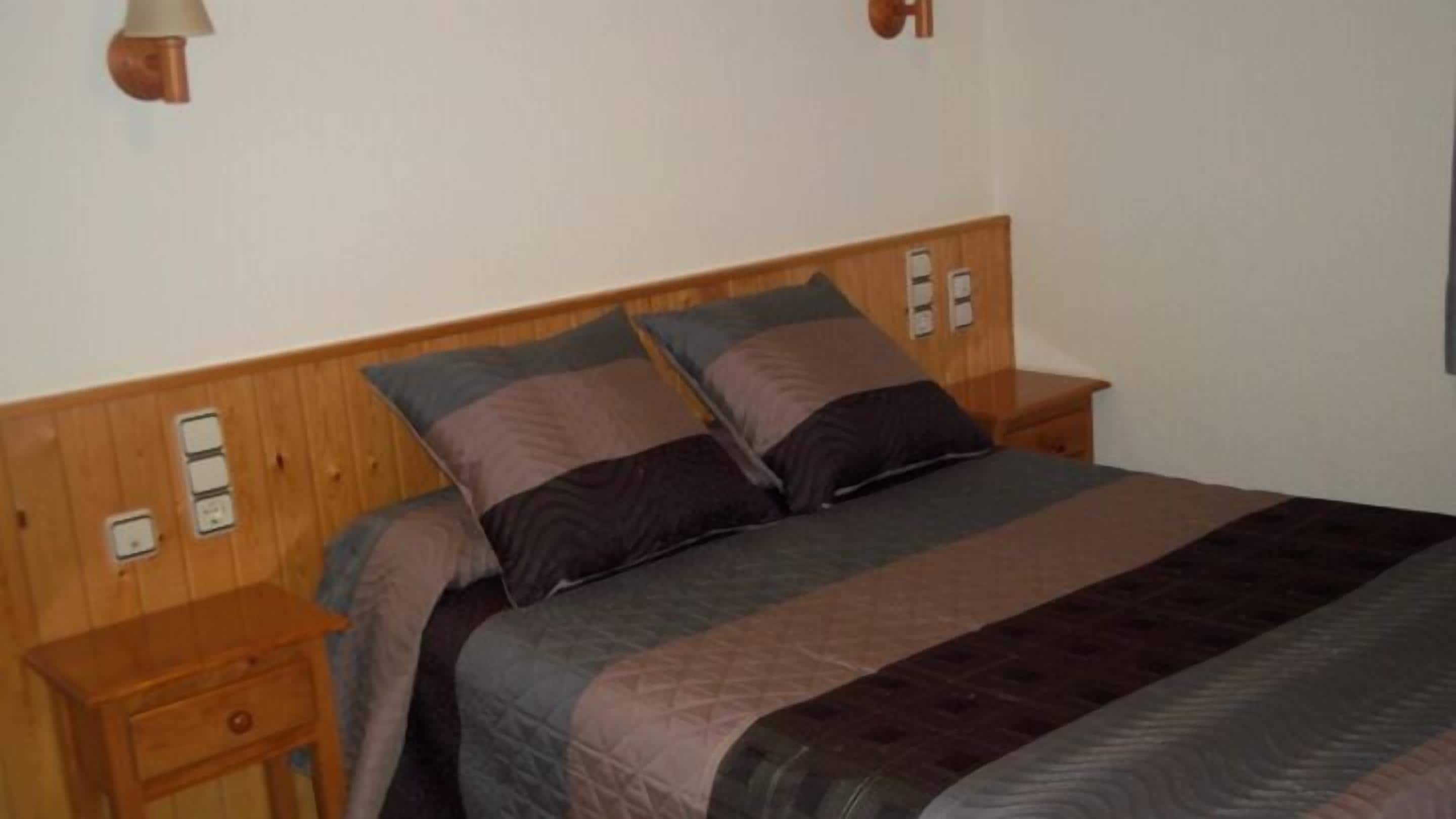 Een slaapkamer met een houten hoofdbord, een bed met meerdere kussens, een nachtkastje en een wandlamp.