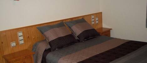 Un dormitorio con cabecera de madera, cama con muchos almohadones, mesita de noche y lámpara fijada a la pared.