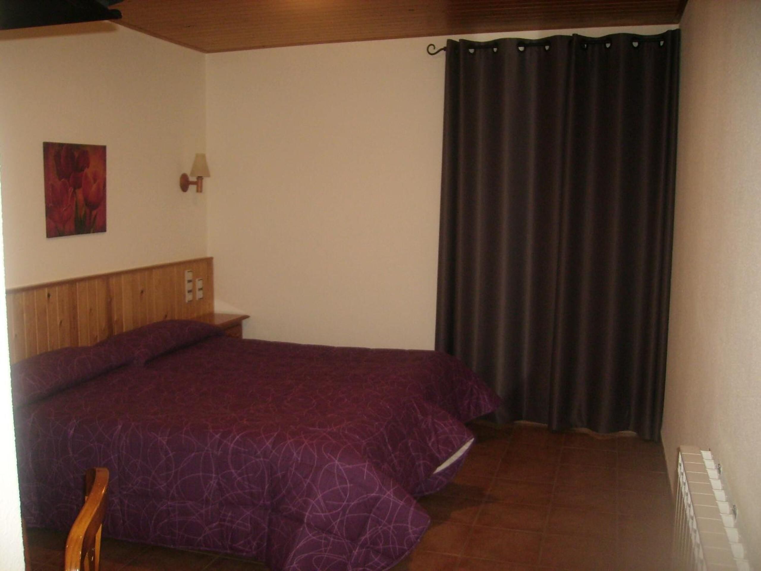 A bedroom with a bed, a wooden headboard, a wall-mounted lamp, and a dark curtain.