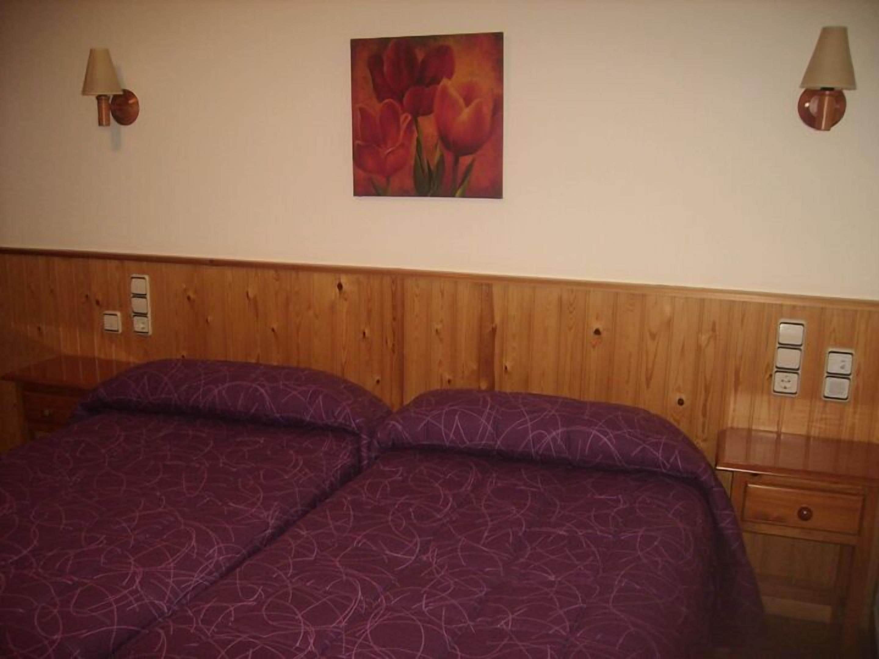 Una cama doble con una colcha morada estampada, un cabecero de madera, dos lámparas fijadas a la pared y un cuadro de tulipanes rojos.