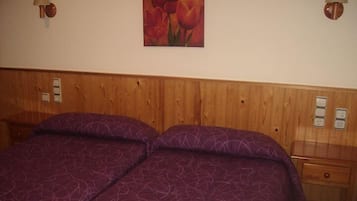 Una cama doble con una colcha morada estampada, un cabecero de madera, dos lámparas fijadas a la pared y un cuadro de tulipanes rojos.