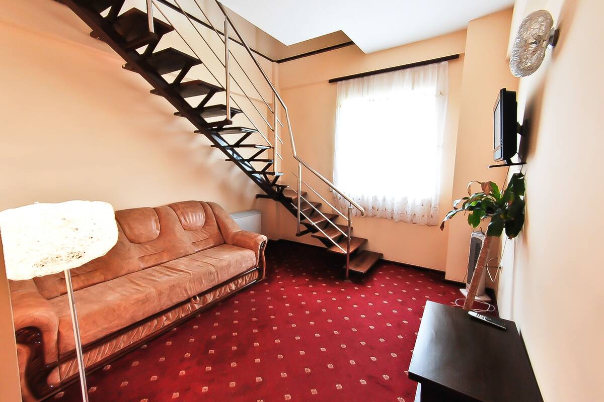 A living room with a brown leather sofa, a red patterned carpet, a black staircase, a white lamp, and a potted plant.