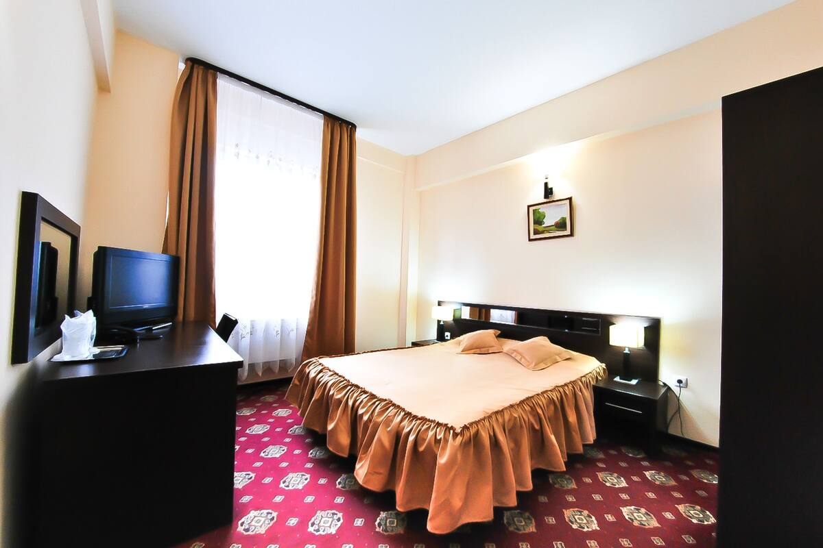 A hotel room with a bed, a desk with a television, and a window with curtains.