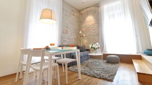 A modern living room with a stone accent wall, a dining table with chairs, a sofa, and a small side table with a lamp and decorative items. - The Bridge Luxury Apartments (Zadar)
