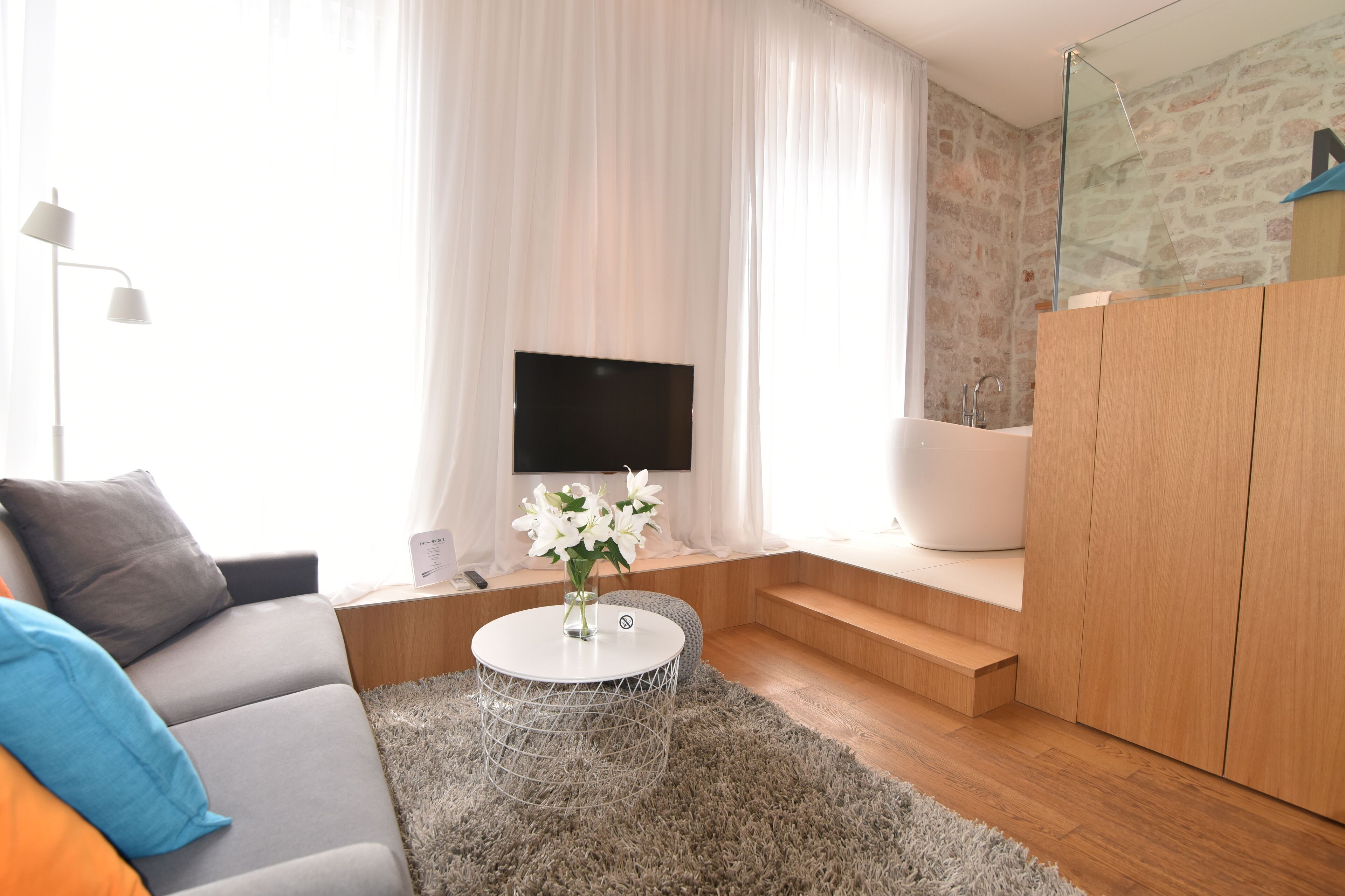 A modern living room with a sofa, a round coffee table with a vase of flowers, a flat-screen TV, a glass-enclosed bathtub, and a stone wall.