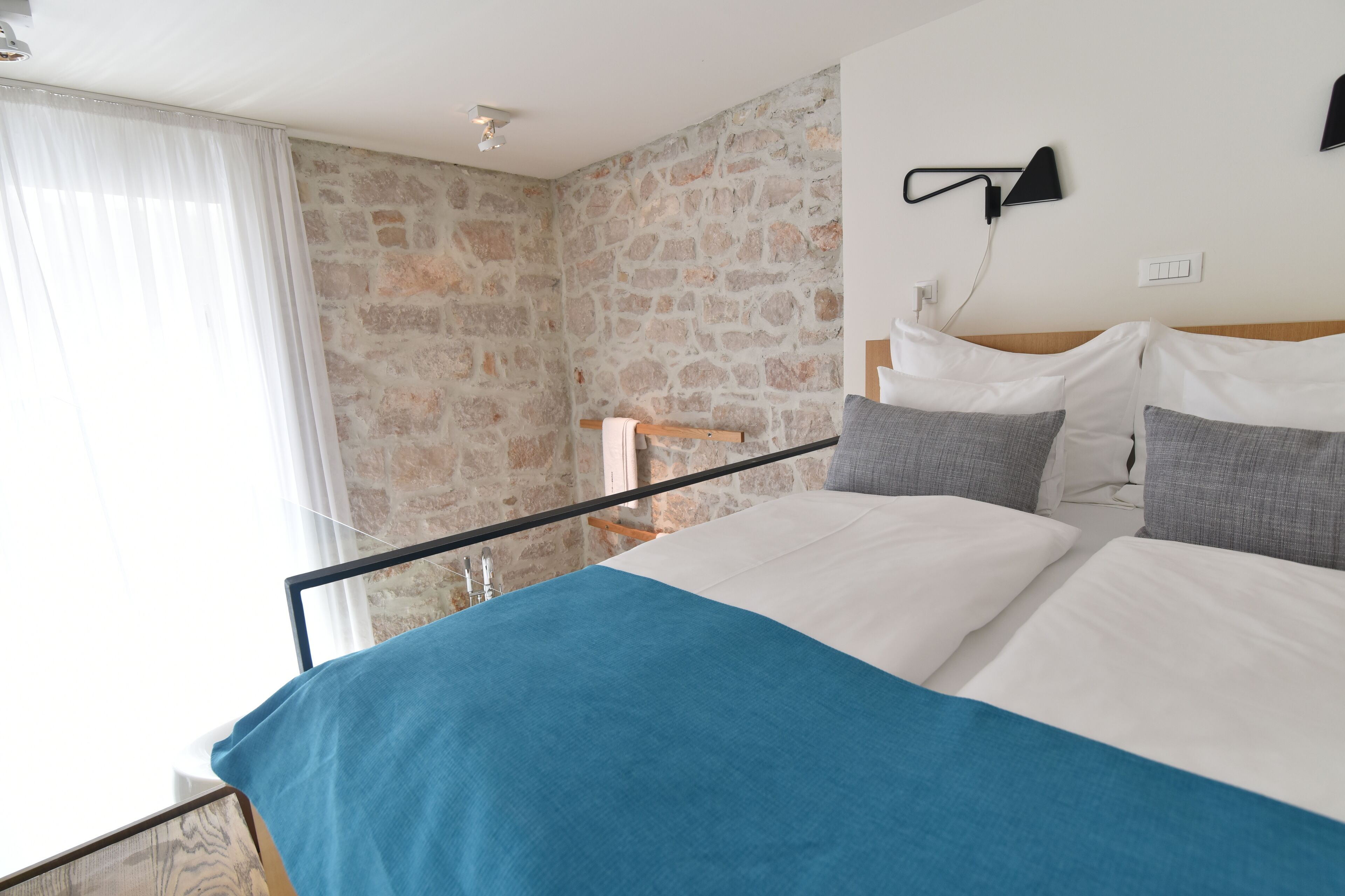 A bedroom with a stone wall, a bed with white and blue bedding, and a nightstand with a lamp.
