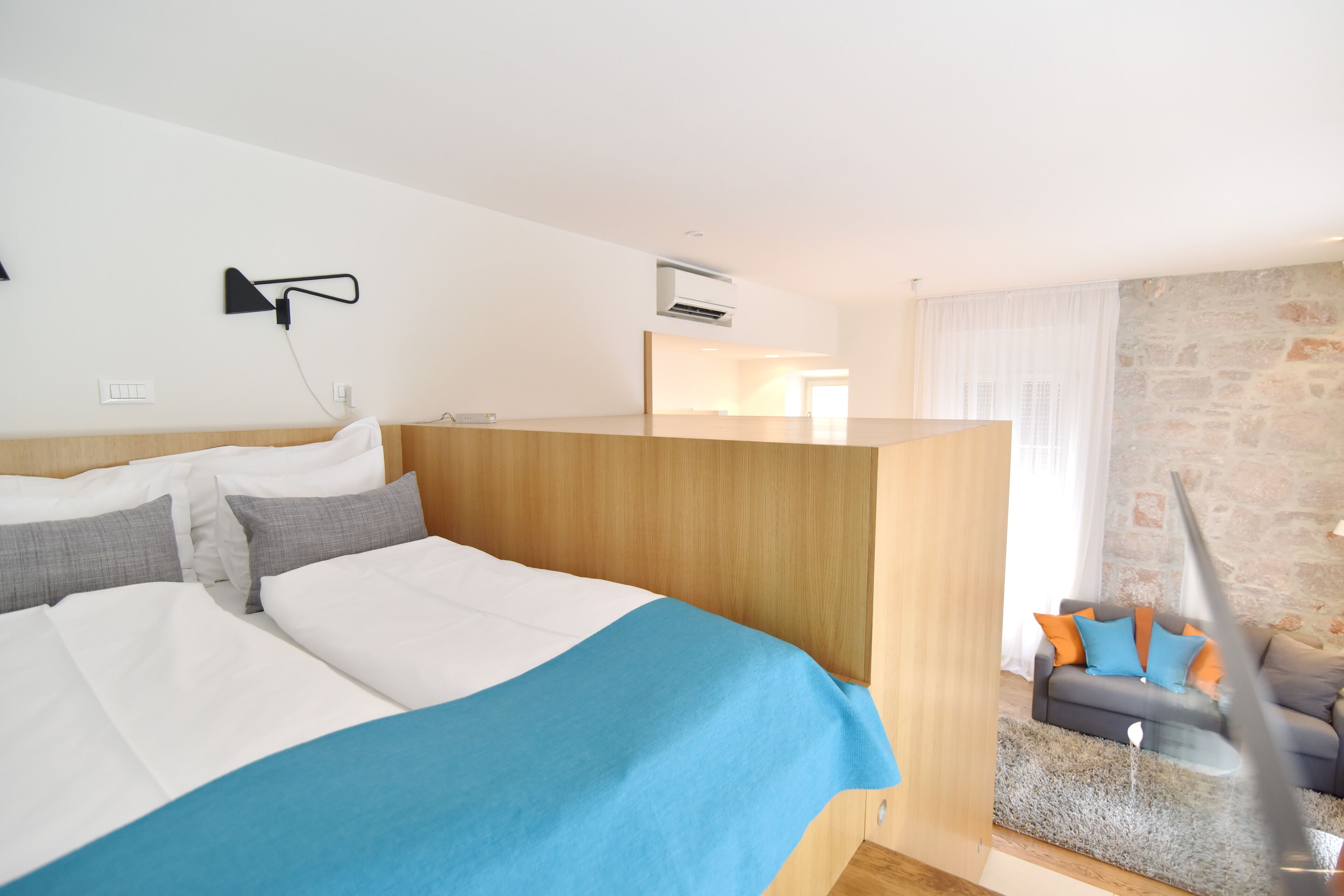 Une chambre d’hôtel moderne avec un grand lit, une tête de lit en bois, un petit coin salon avec un canapé et un mur en pierre.