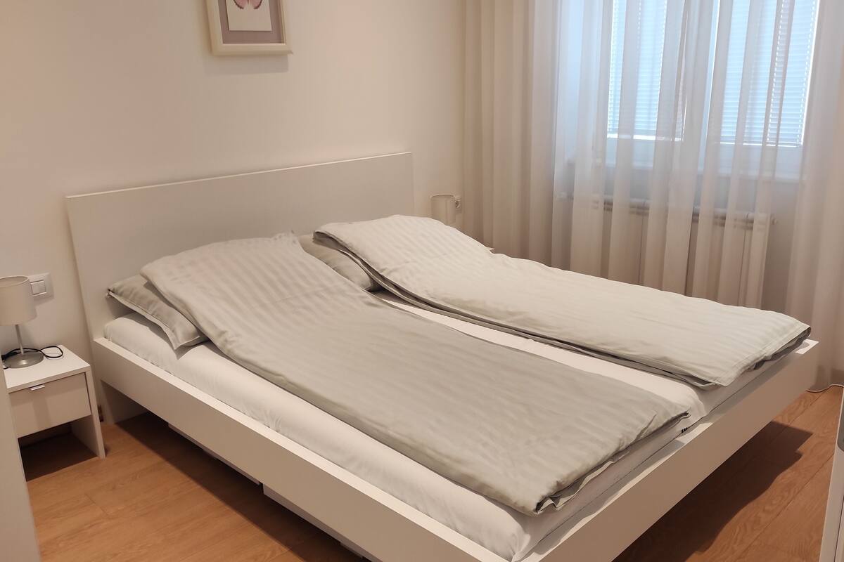 Ein Doppelbett mit Kopfteil, ein Nachttisch mit Lampe sowie ein Fenster mit durchscheinenden Vorhängen.