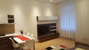 Un salon moderne comprenant un coin repas, un téléviseur à écran plat et une grande fenêtre ornée de rideaux légers.
