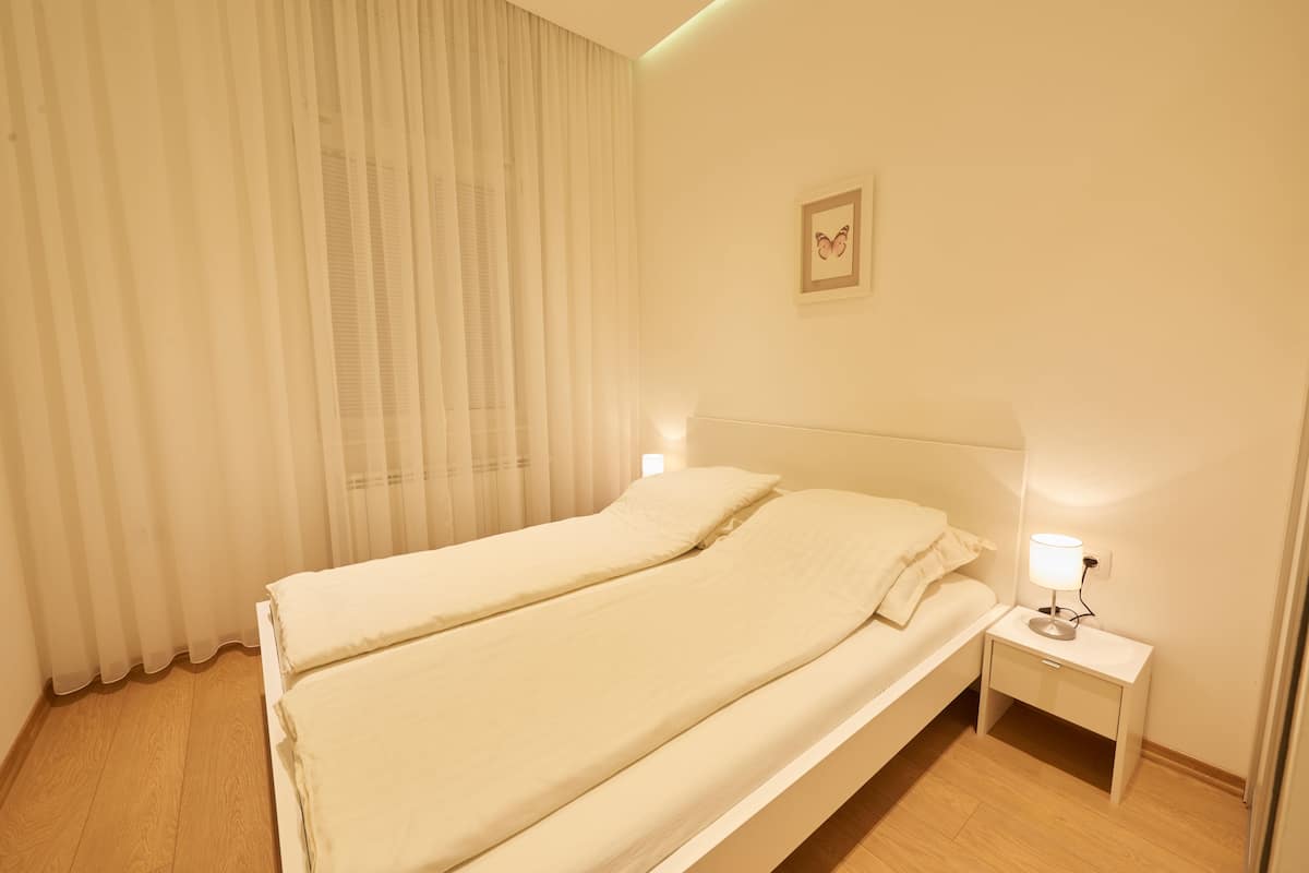 Ein ordentlich gemachtes Bett mit weißer Decke und Kissen, ein Nachttisch mit einer Lampe sowie ein an der Wand hängendes Bild.