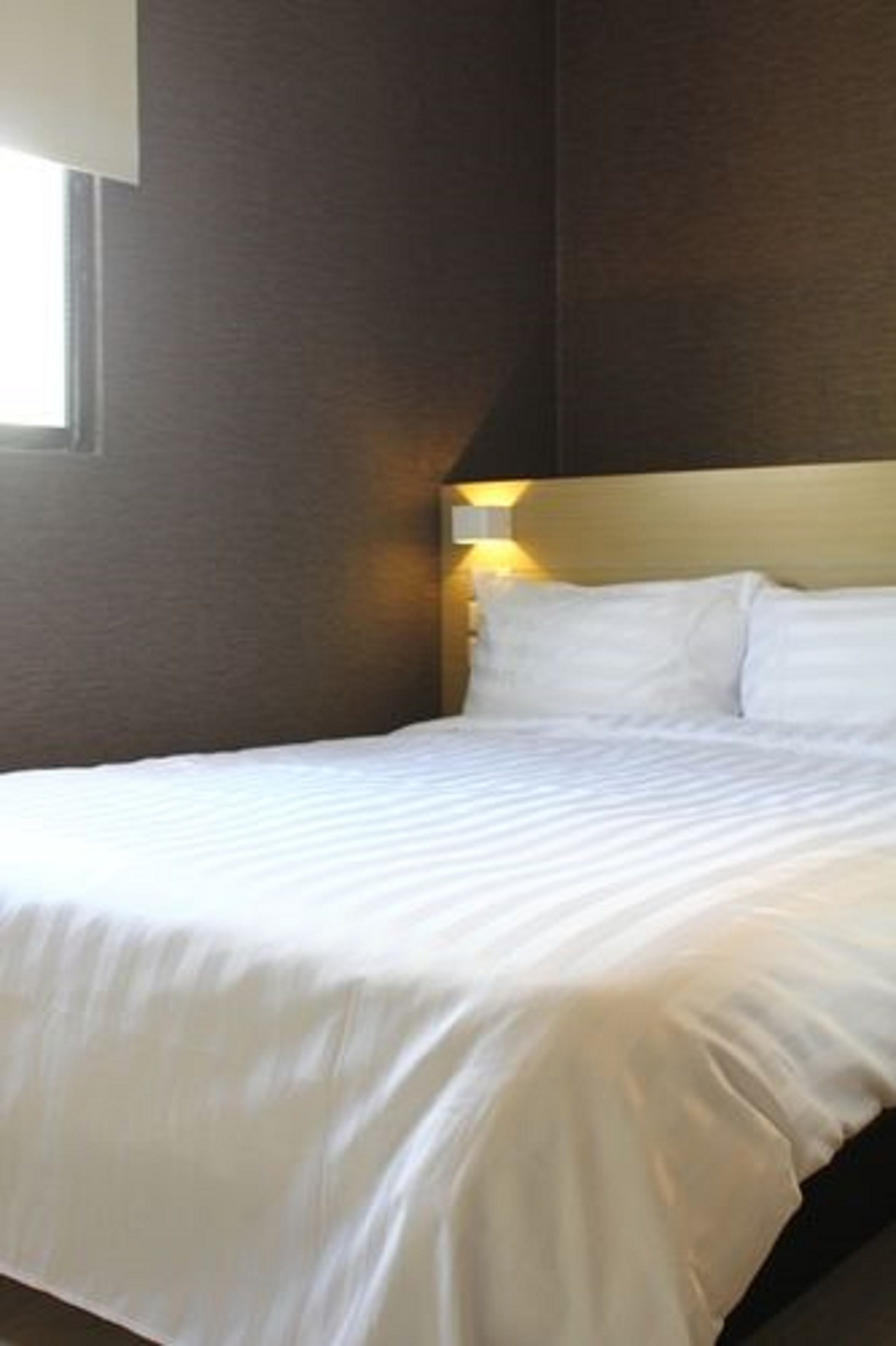 A neatly made bed in a hotel room with a headboard, a bedside lamp, and a window.