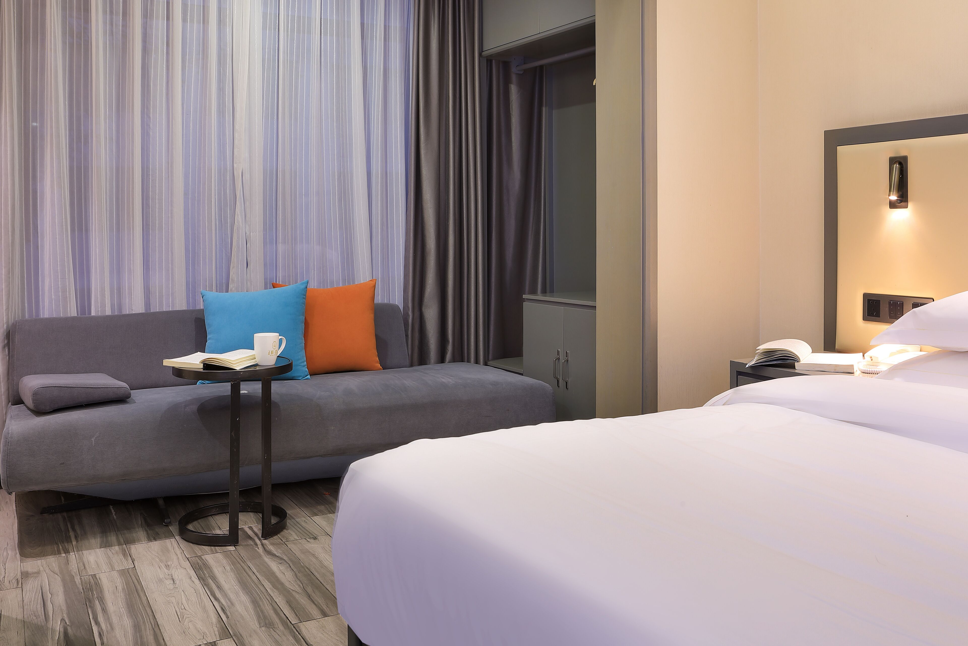 Ein Hotelzimmer mit grauem Sofa, einem kleinen Tisch mit Buch und Tasse, einem weißen Bett und einem Fenster mit Vorhängen.