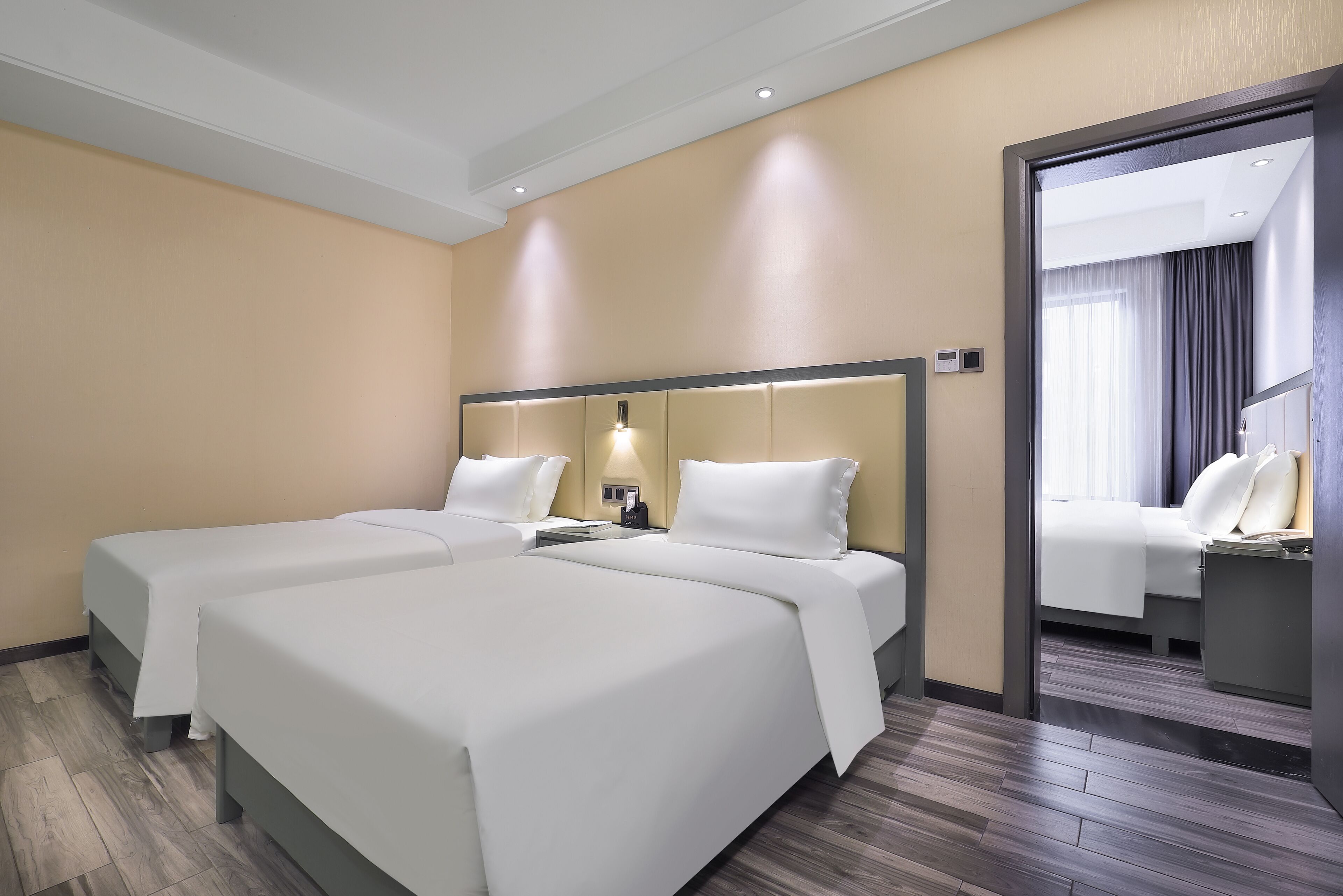A hotel room with two beds, a wooden floor, a beige wall, and a mirror.
