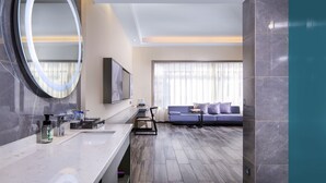 Un baño moderno con un espejo grande, un lavabo y una cómoda. En la zona de estar contigua hay un sofá y un televisor.
