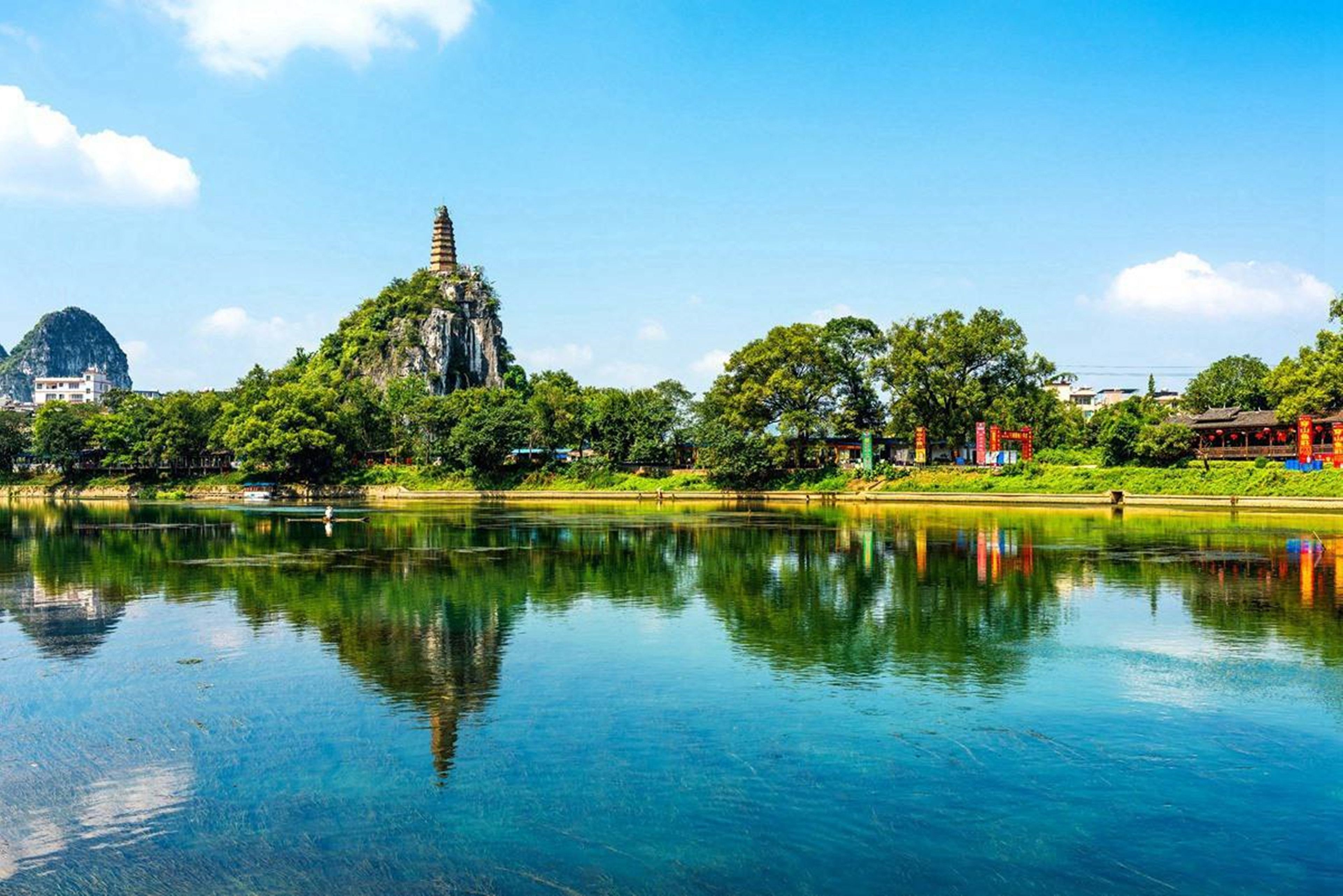 A serene lake with a temple on a hill, surrounded by lush greenery and a clear sky.