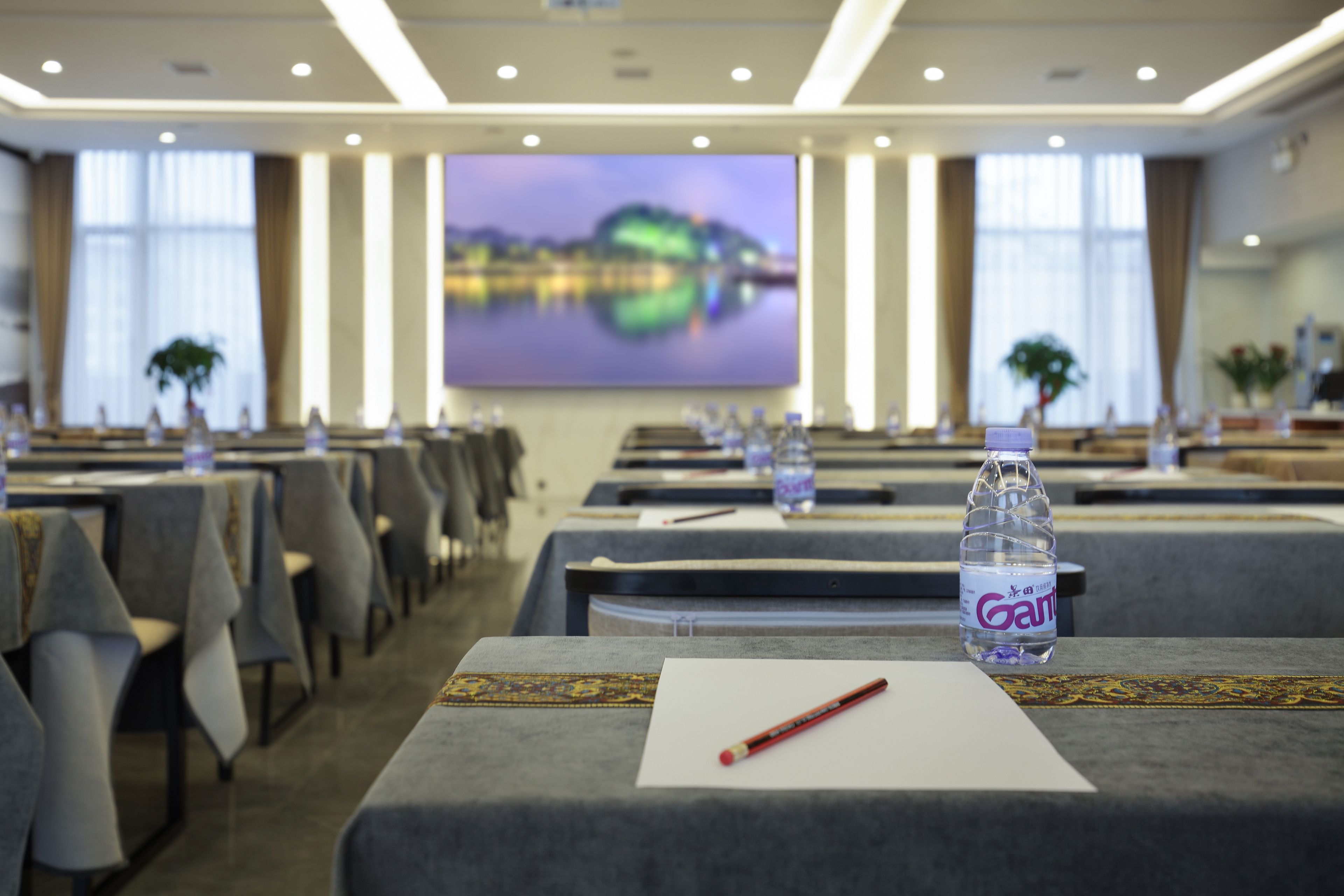 Un salón de conferencias con filas de mesas, una pantalla de proyector y una botella de agua sobre una de ellas.